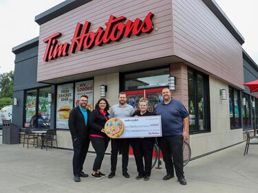 Owners of Tim Hortons stand in front of Tim Hortons holding large cheque
