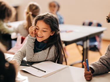  girl laughs happily in a primary school class, she is sitting at a table with her her classmates.