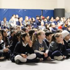 Students sitting on floor, parents sitting on chairs behind them looking at a screen of a video playing